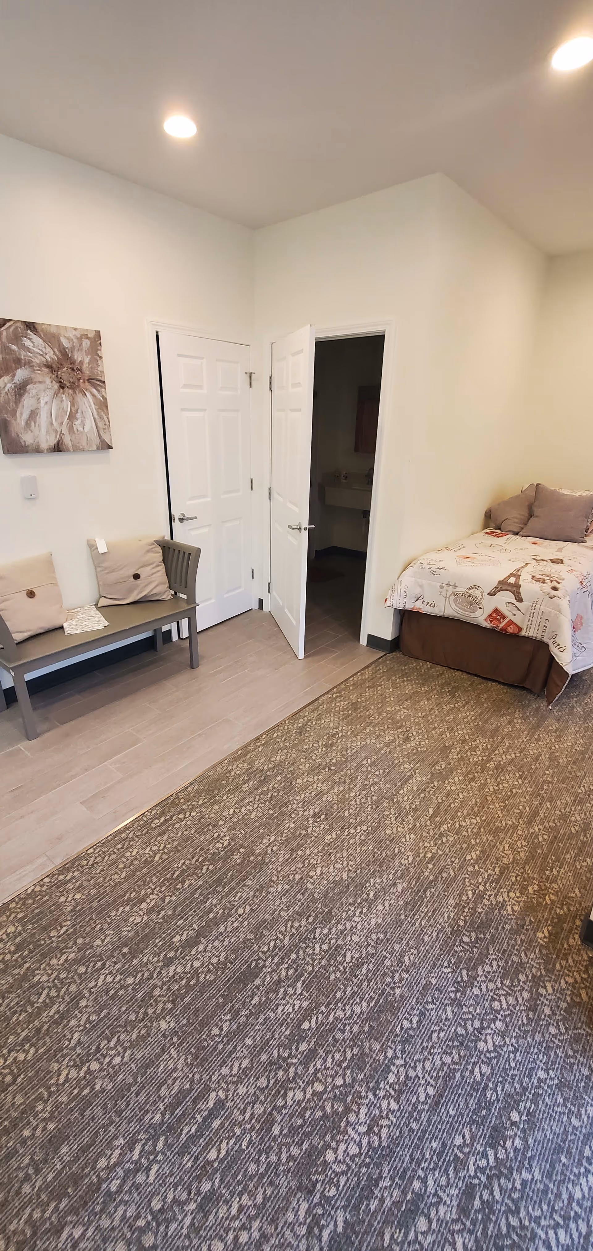 A small bedroom with a single bed covered in a Paris-themed bedspread, a bench with two cushions against the wall, a floral painting above the bench, and two white doors, one slightly open revealing a sink inside.