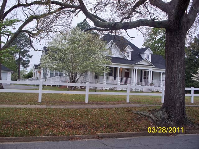 A large white house with a wraparound porch and a dark roof, surrounded by trees and a white fence. The lawn is covered with fallen leaves and there is a large tree in the foreground. The date 03/28/2011 is visible in the bottom right corner.