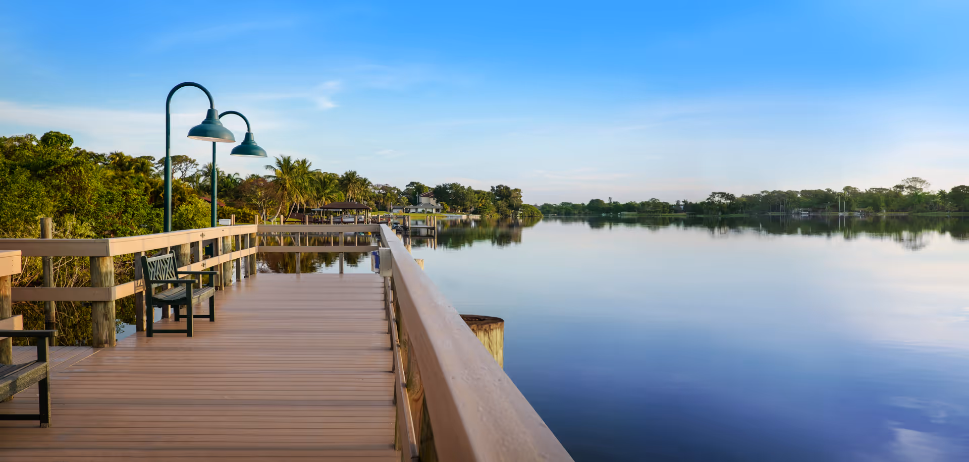 A wooden lakeside boardwalk with benches and lamp posts overlooking a calm reflective lake and tree-lined shore under a blue sky.