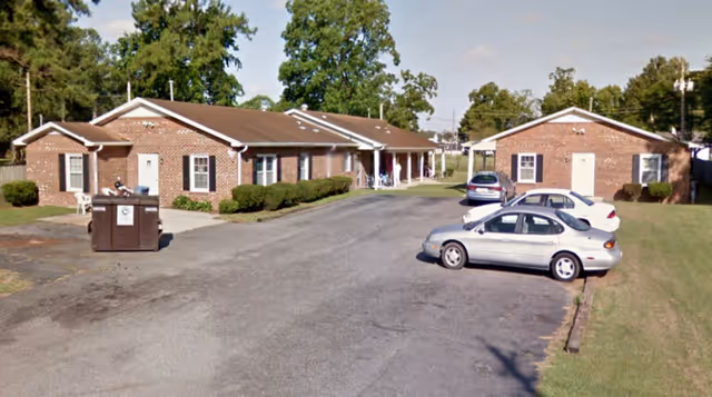 View of a senior living facility parking area with two single-story brick buildings on either side. There are three cars parked on the asphalt driveway and a large dumpster near the left building. The area is surrounded by grass and trees under a partly cloudy sky.