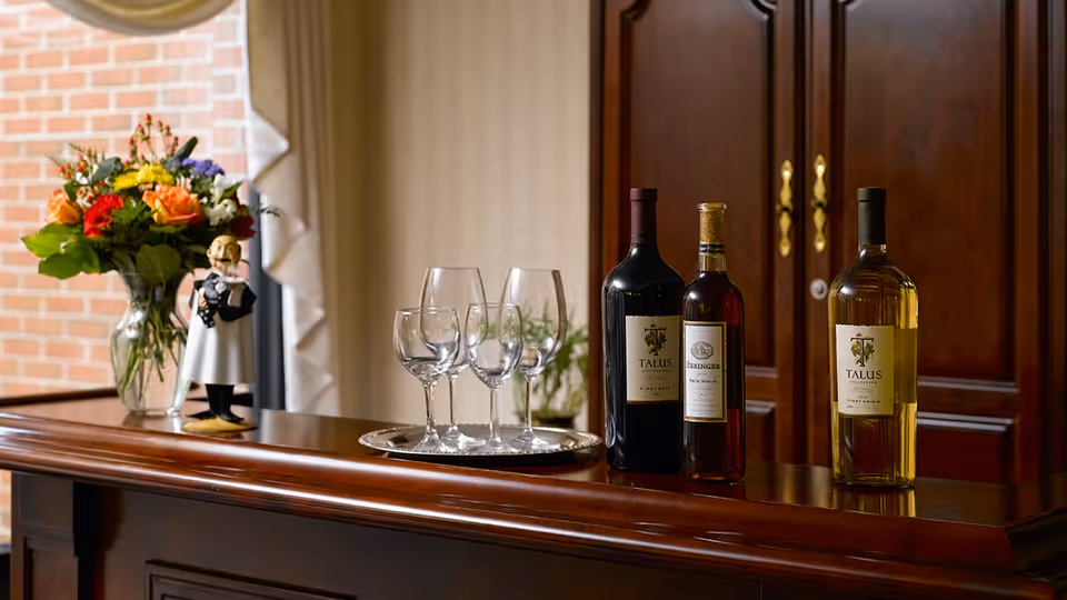 A polished wooden bar counter with three bottles of wine, a silver tray holding four empty wine glasses, a vase of colorful flowers, and a small figurine of a waiter. Behind the counter is a wooden cabinet with brass handles and a window with a brick wall visible outside.