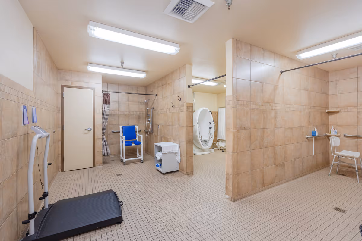 A spacious, tiled bathroom area in a senior care facility with multiple accessible shower stations. The room features beige wall and floor tiles, a blue shower chair, grab bars, a treadmill, and a specialized bathing tub in the background. The lighting is bright with ceiling-mounted fluorescent lights.