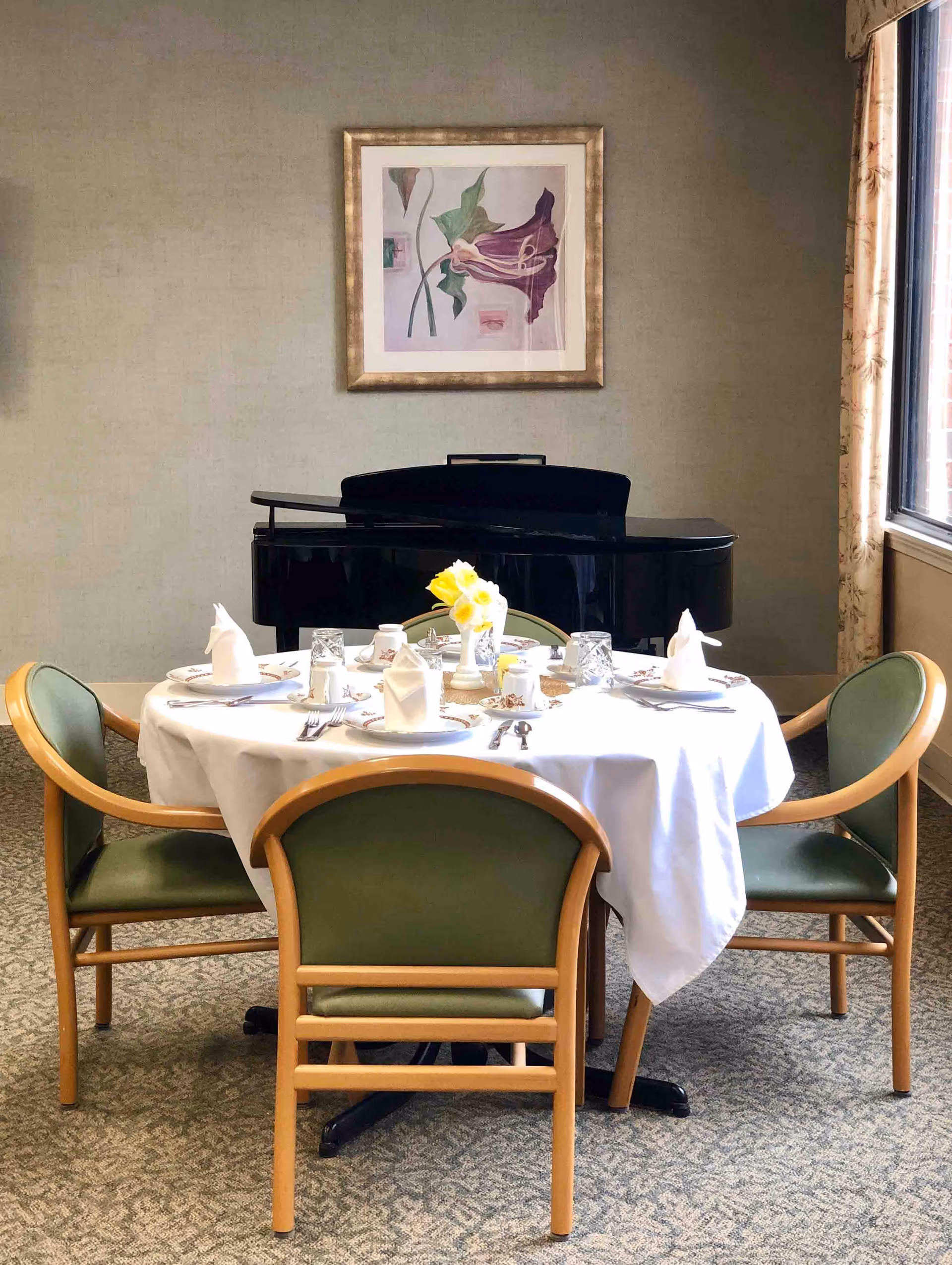 A round dining table set with white tablecloth, plates, glasses, silverware, and napkins folded in a decorative manner. A vase with yellow flowers is in the center of the table. Four green cushioned chairs with wooden frames surround the table. In the background, there is a black piano and a framed floral artwork on the wall. A window with floral curtains is on the right side, allowing natural light into the room.