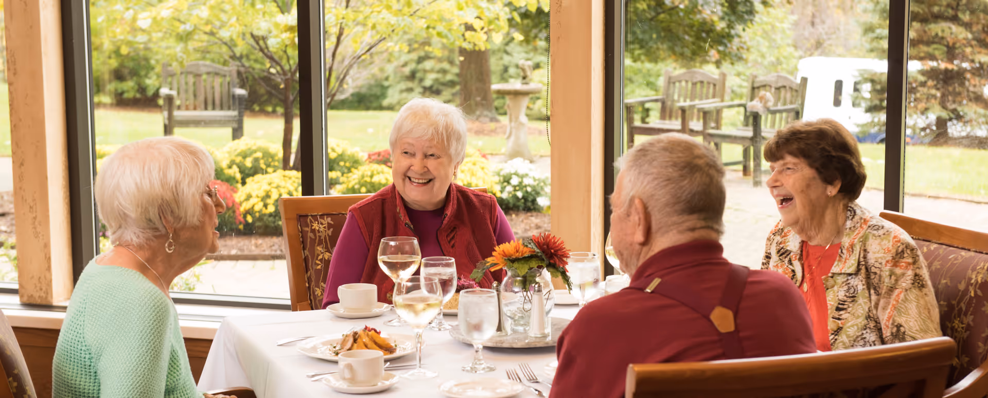 Four elderly people sitting around a dining table inside a senior living facility, smiling and enjoying a meal together. The table is set with plates, glasses of water and wine, and a small floral centerpiece. Large windows behind them show a garden with trees, flowers, and benches outside.