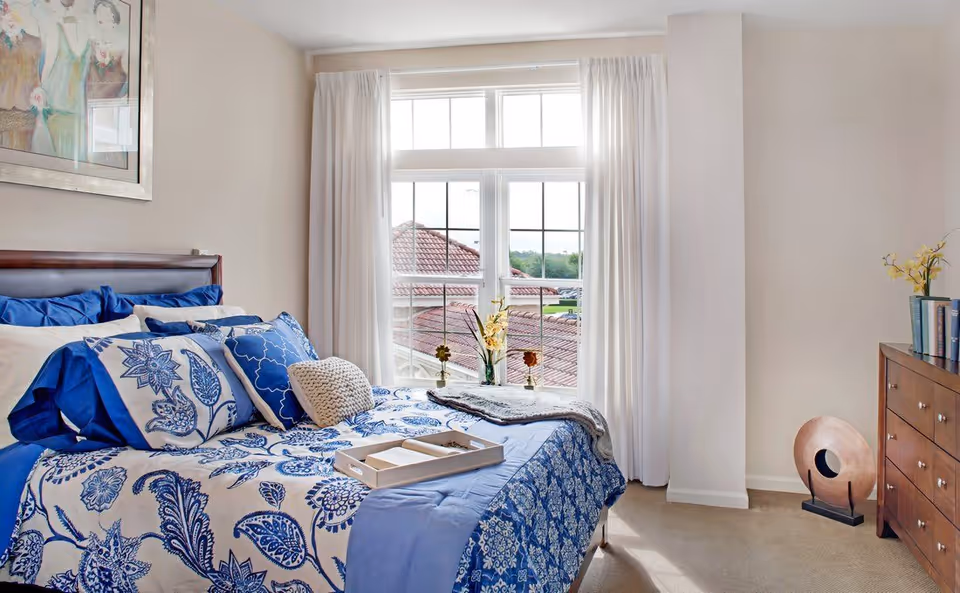 A bright bedroom with a large window covered by white curtains. The bed has a blue and white floral patterned comforter and multiple pillows in shades of blue and white. A tray with books is placed on the bed. There is a wooden dresser on the right side with books and a vase with yellow flowers. A decorative circular sculpture is on the floor next to the dresser. A framed painting hangs on the wall above the bed.