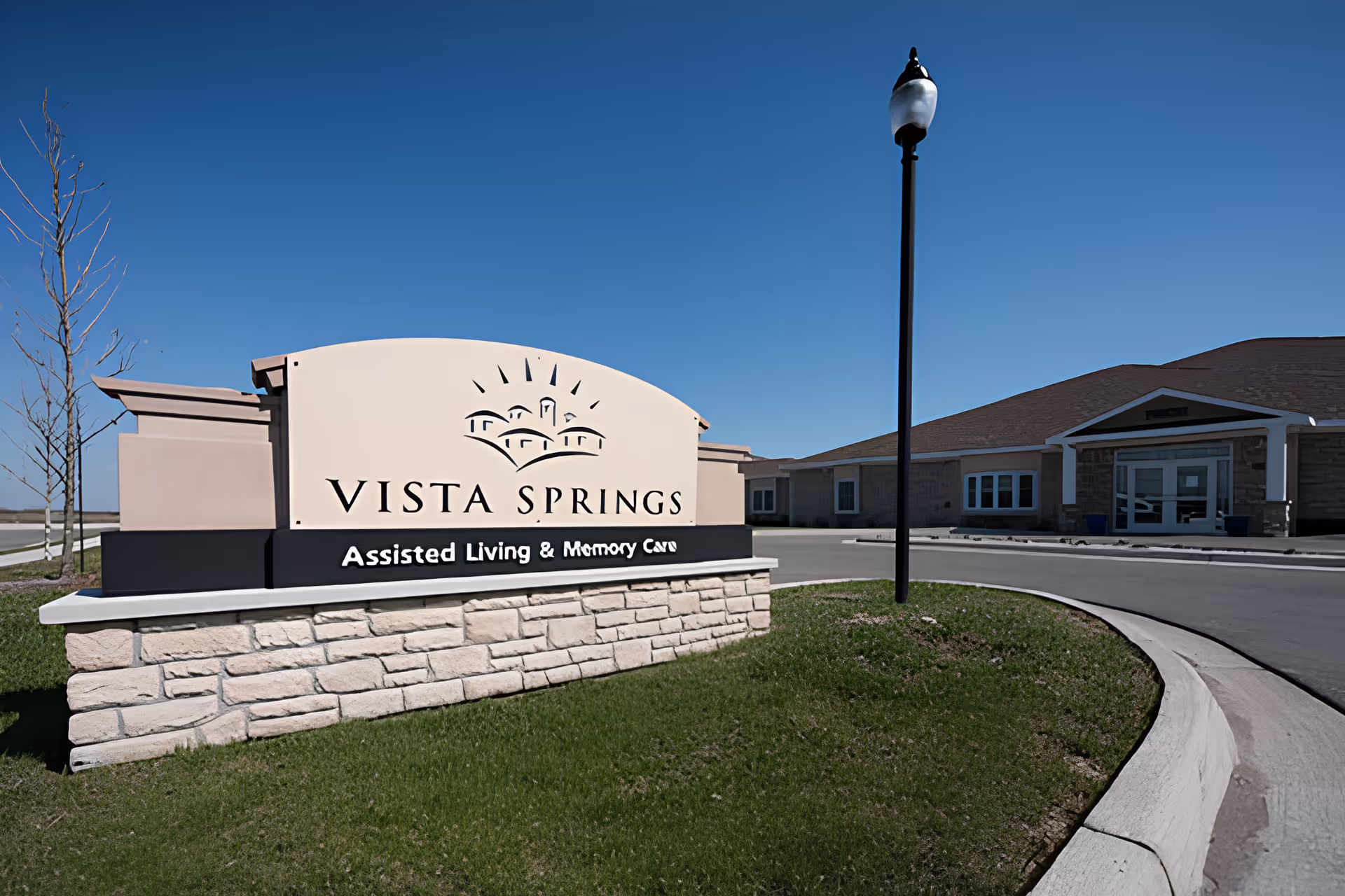 Outdoor view of the entrance area of Vista Springs Assisted Living & Memory Care facility, featuring a large sign with the facility name and a lamp post near the driveway under a clear blue sky.