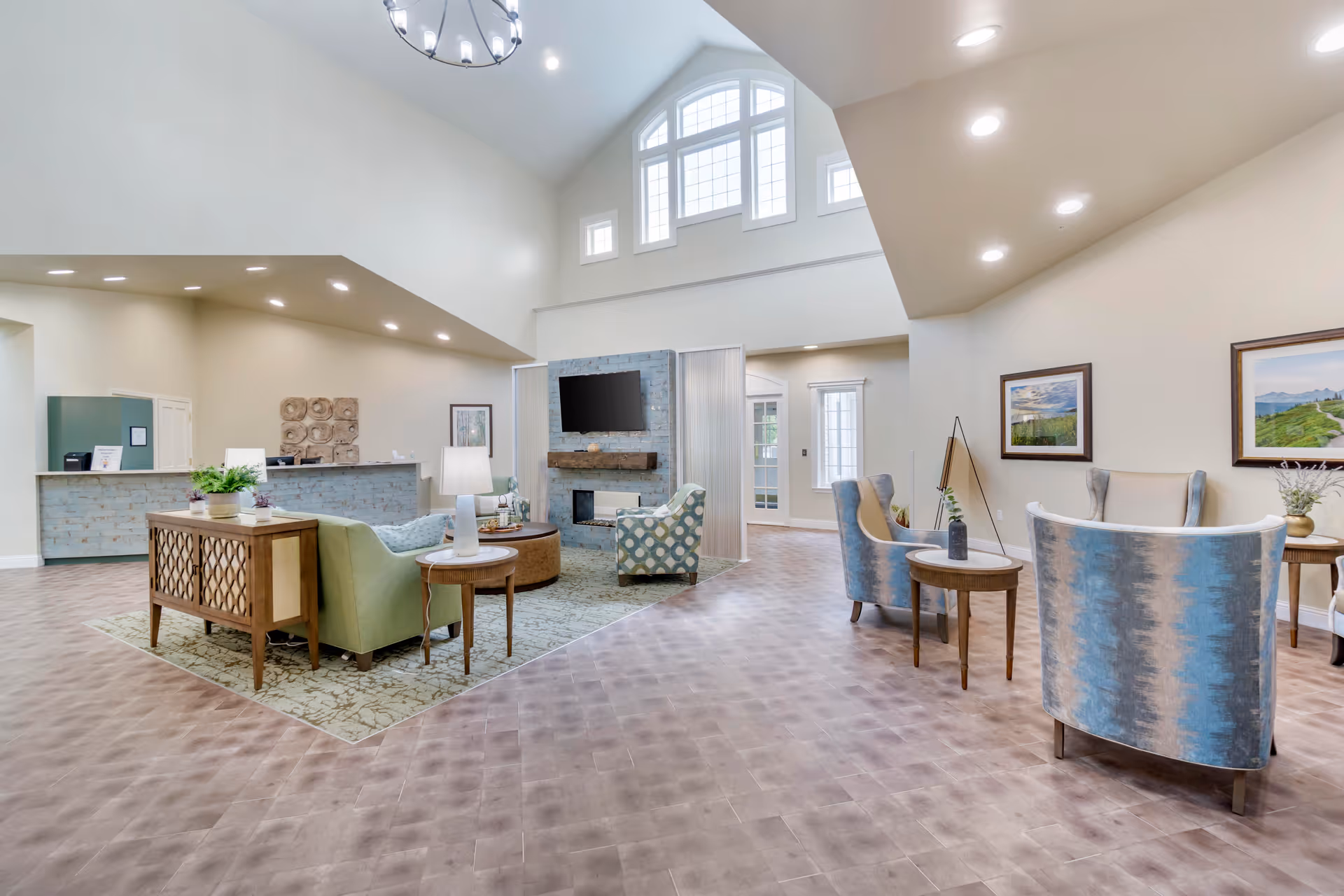 A spacious and well-lit living room area in a senior living facility with high ceilings and large windows. The room features a seating arrangement with a green sofa, patterned armchairs, round wooden tables, and a TV mounted above a stone fireplace. The walls are decorated with framed landscape paintings, and there is a reception desk visible in the background.