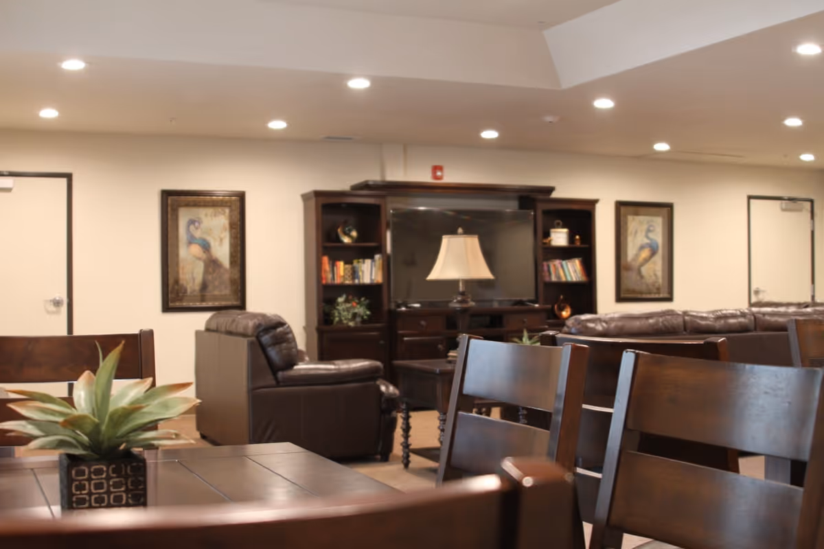 A senior living facility common area with dark wooden chairs and tables in the foreground, a leather armchair and sofa, a large flat-screen TV on a wooden entertainment center, two framed peacock paintings on the wall, and a table lamp in the center.