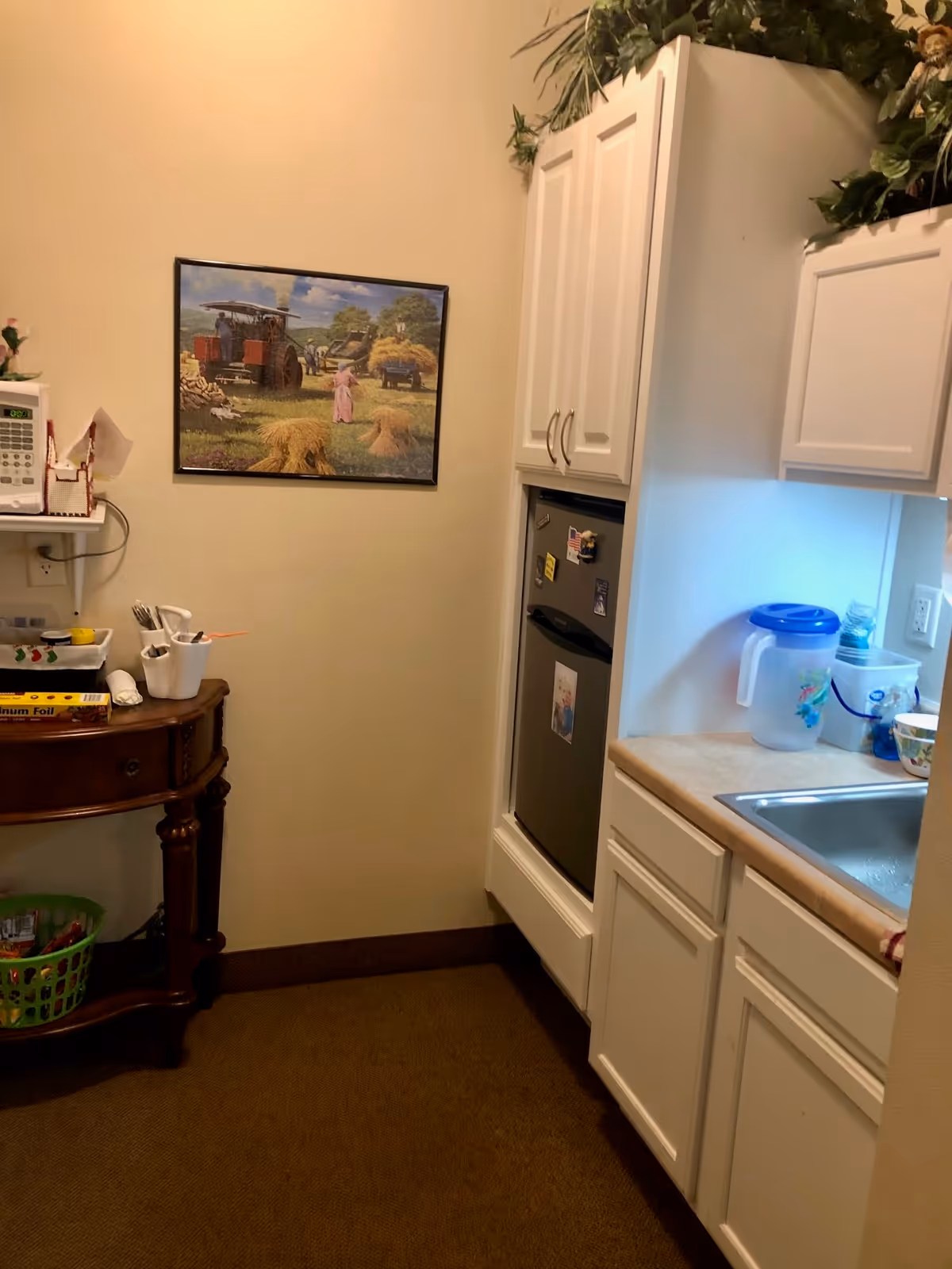 Small kitchenette with white cabinets, a sink and mini refrigerator, countertop items, a wooden side table and framed artwork on the wall.