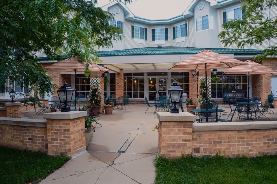 Outdoor patio area of a senior living facility with several round metal tables and chairs, each shaded by large striped umbrellas. The patio is surrounded by a low brick wall with lantern-style lights on the pillars. There are potted plants and greenery around the space, and the building's exterior with large windows and a green roof is visible in the background.