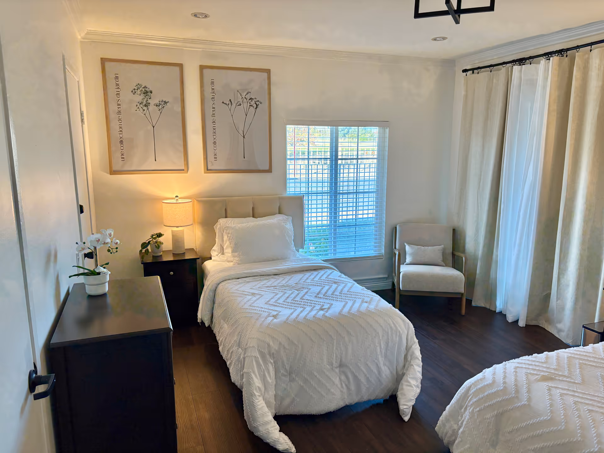 Bright bedroom with two twin beds, a nightstand and lamp, dresser, armchair, and a window with curtains.