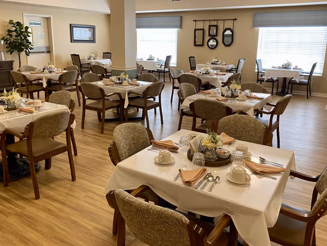 A bright dining room with multiple neatly set tables and upholstered chairs arranged on wood-look flooring.