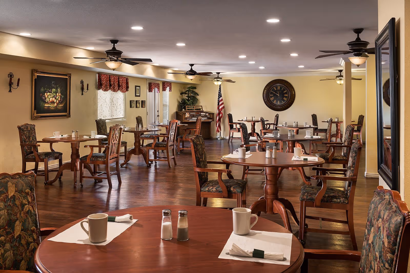 A spacious dining room with multiple round wooden tables and cushioned chairs arranged neatly. Each table has a white placemat, a coffee mug, and salt and pepper shakers. The room features ceiling fans with lights, a large wall clock, an American flag, framed artwork, and a piano in the background. The walls are painted a warm beige color, and the floor is polished wood.