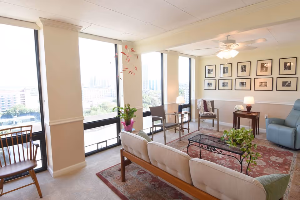 A bright and cozy living room area with large windows offering a city view. The room features a beige cushioned sofa, several chairs, a glass-top coffee table with a plant, a side table with a lamp and flowers, a ceiling fan with lights, and framed black-and-white photographs on the wall. The floor is carpeted with a red patterned rug.