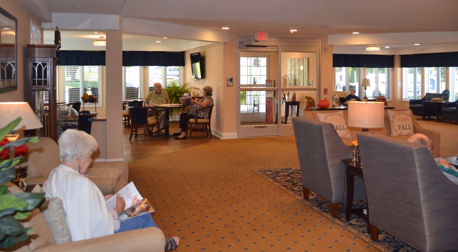 A spacious senior living facility common area with several elderly residents seated and engaged in various activities. The room features comfortable chairs, a carpeted floor, large windows with blue valances, and a glass door entrance. There are tables with chairs near the windows, and the decor includes lamps, plants, and seasonal fall-themed pillows.