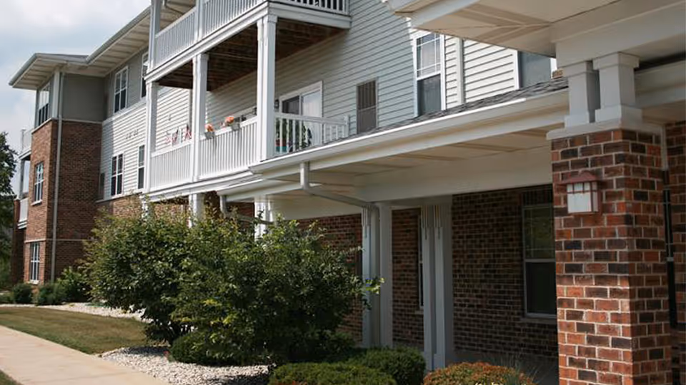 Exterior view of a senior apartment building with brick and light-colored siding, featuring balconies with railings and some potted plants. There are bushes and a sidewalk in front of the building under a partly cloudy sky.
