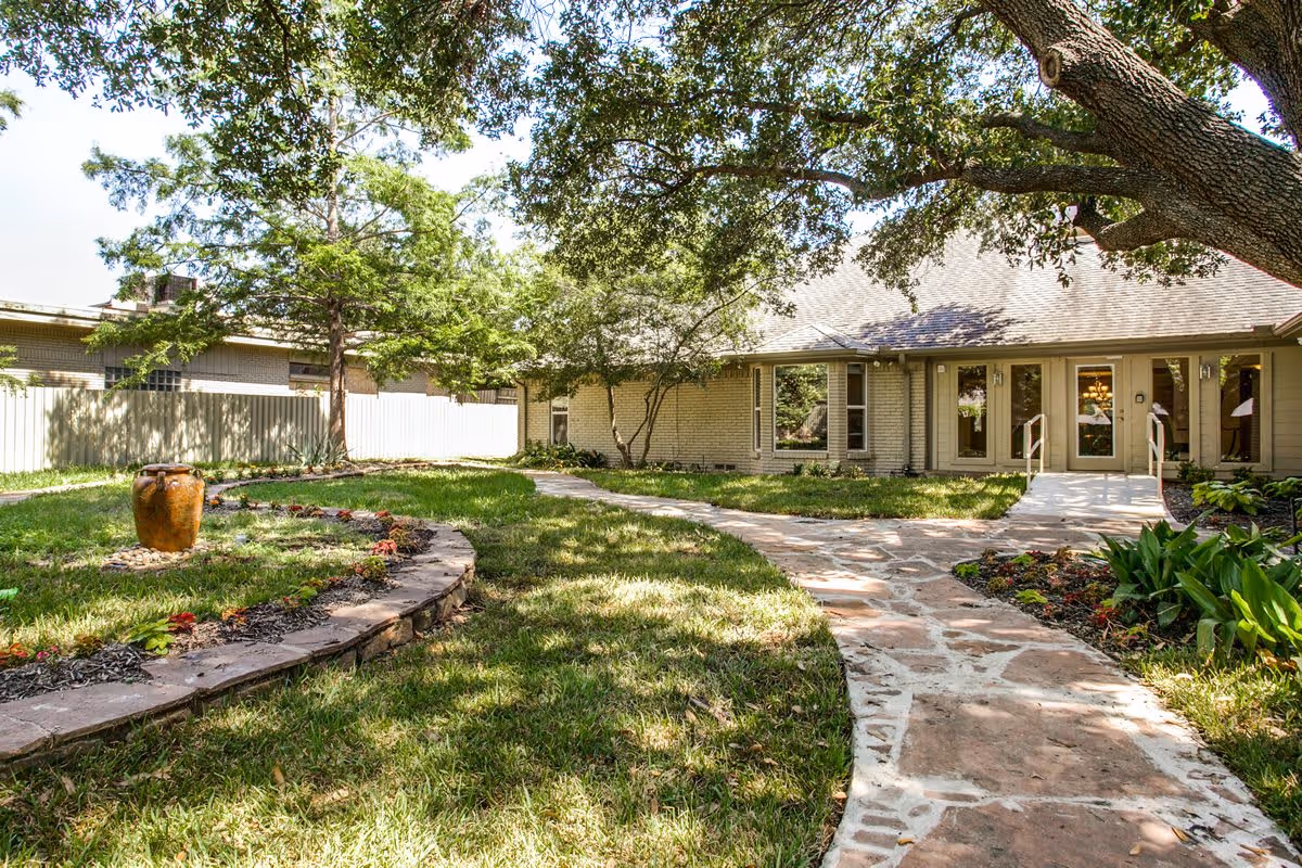 A peaceful outdoor garden area with a stone pathway leading to the entrance of a single-story building. The garden features a circular raised flower bed with a large decorative urn in the center, surrounded by green grass, trees, and various plants. The building has large windows and double glass doors, with a shaded area provided by a large tree.