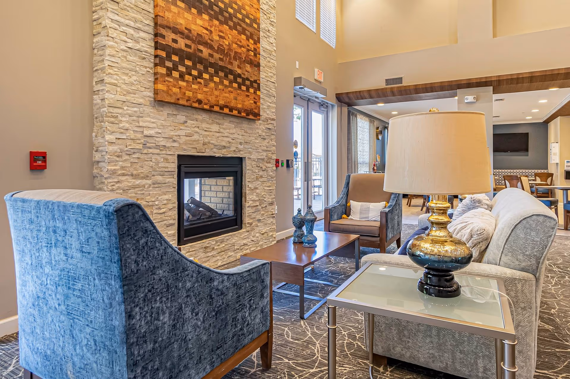A cozy living room area in a senior living facility featuring a stone fireplace with a wooden art piece above it, a blue upholstered armchair, a beige armchair with a pillow, a light gray sofa with pillows, a glass side table with a gold and black lamp, and a wooden coffee table with decorative items. The room has high ceilings, large windows, and an open doorway leading to another seating area with tables and chairs.