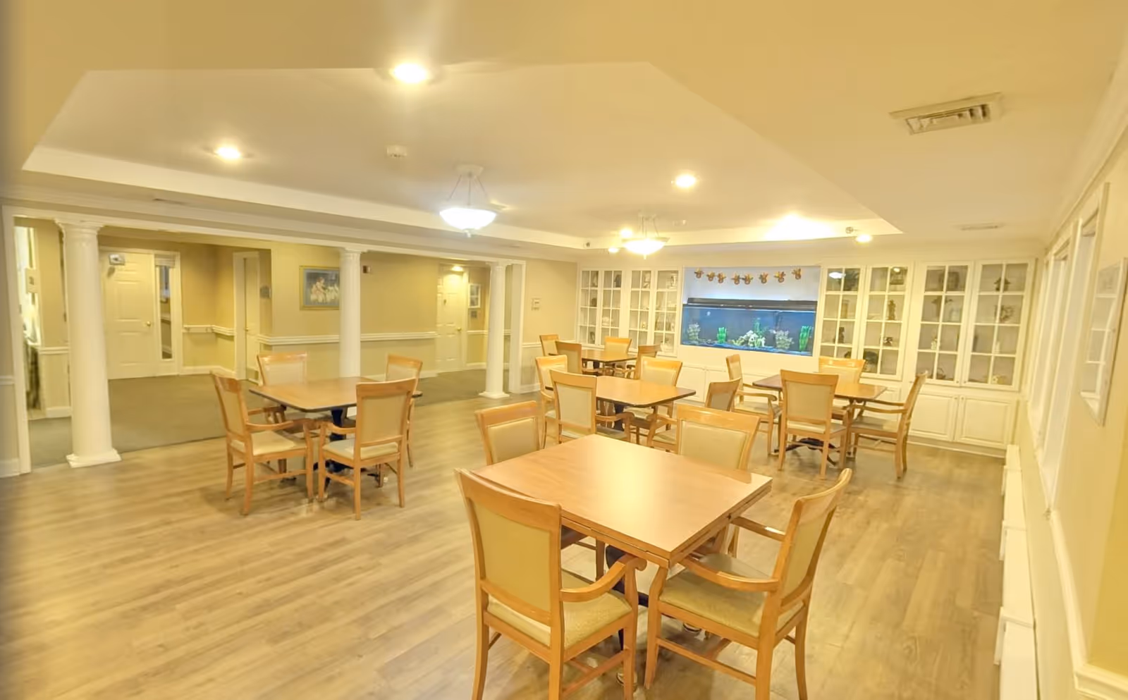 A spacious dining room with multiple wooden tables and chairs arranged neatly on a wood floor. The room features white columns, built-in white cabinets with glass doors, and a large aquarium embedded in the wall. The ceiling has recessed lighting and hanging light fixtures, and the walls are painted a soft yellow color.