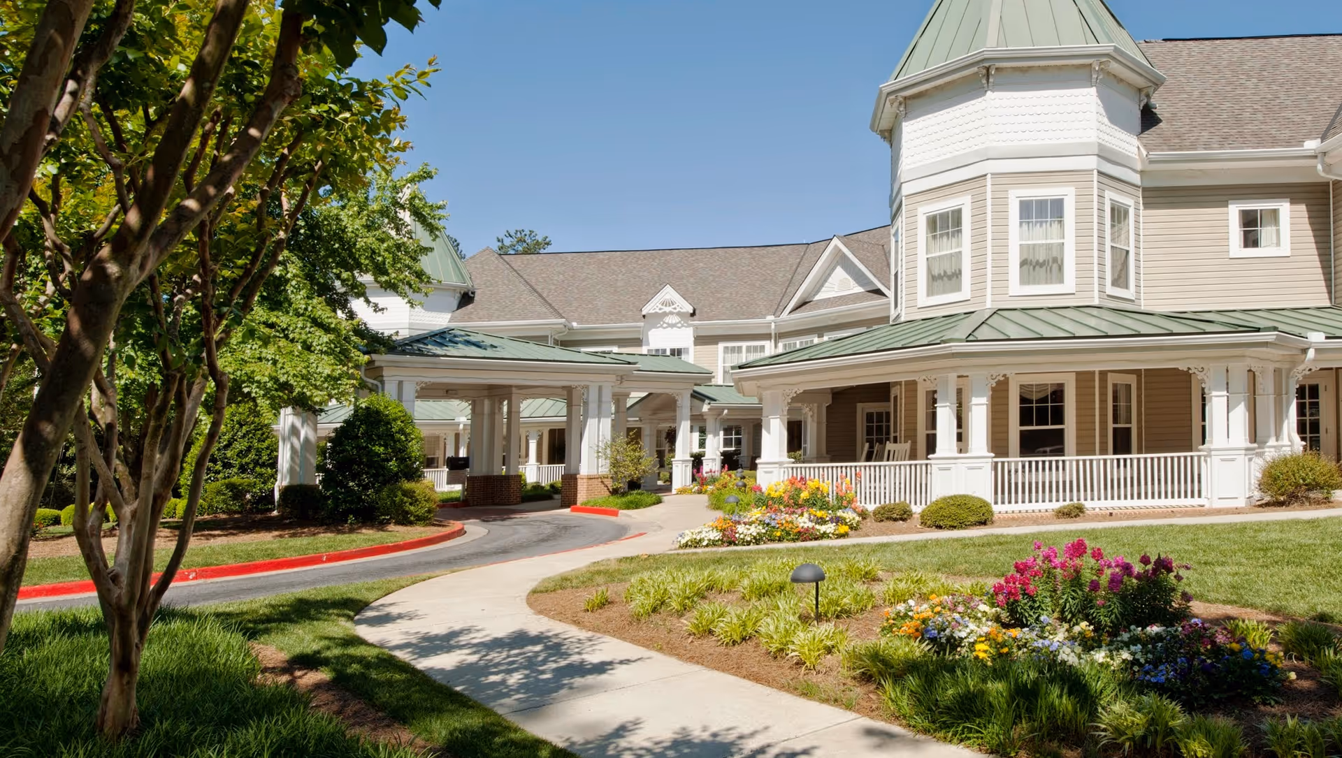 Exterior view of Ivey Ridge Senior Living facility showing a large building with a green metal roof, white columns, and a wrap-around porch. The foreground features a curved sidewalk, well-maintained landscaping with colorful flowers, bushes, and trees under a clear blue sky.