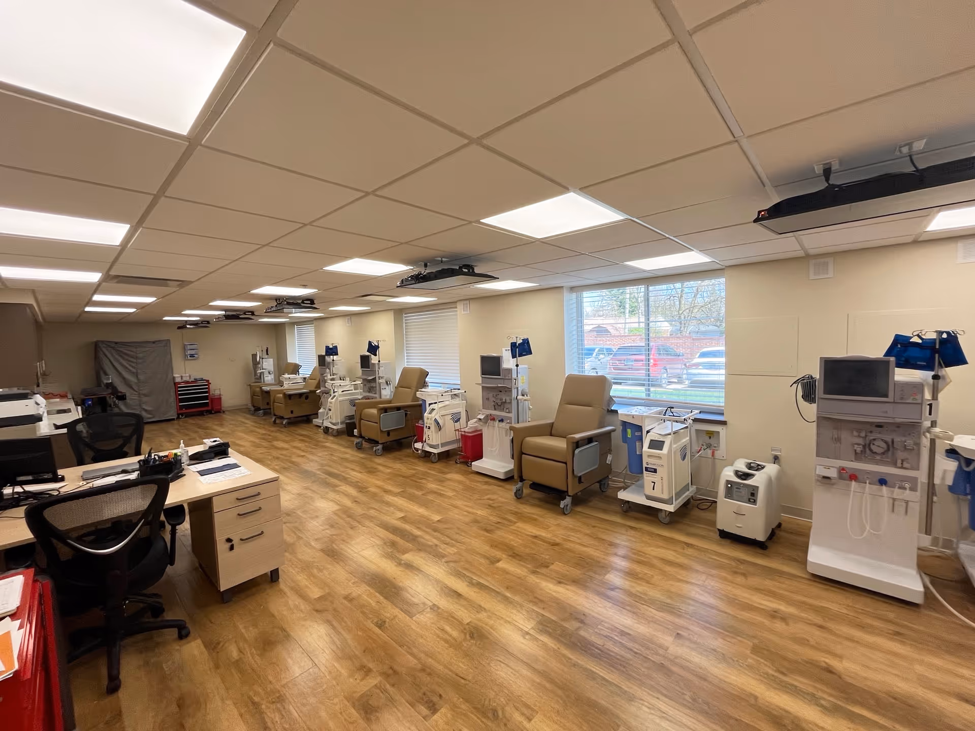 A medical treatment room with multiple dialysis chairs and dialysis machines lined up along the wall under large windows. The room has wood flooring, beige walls, and a drop ceiling with fluorescent lights. There is a desk with office chairs and computer equipment on the left side of the room.