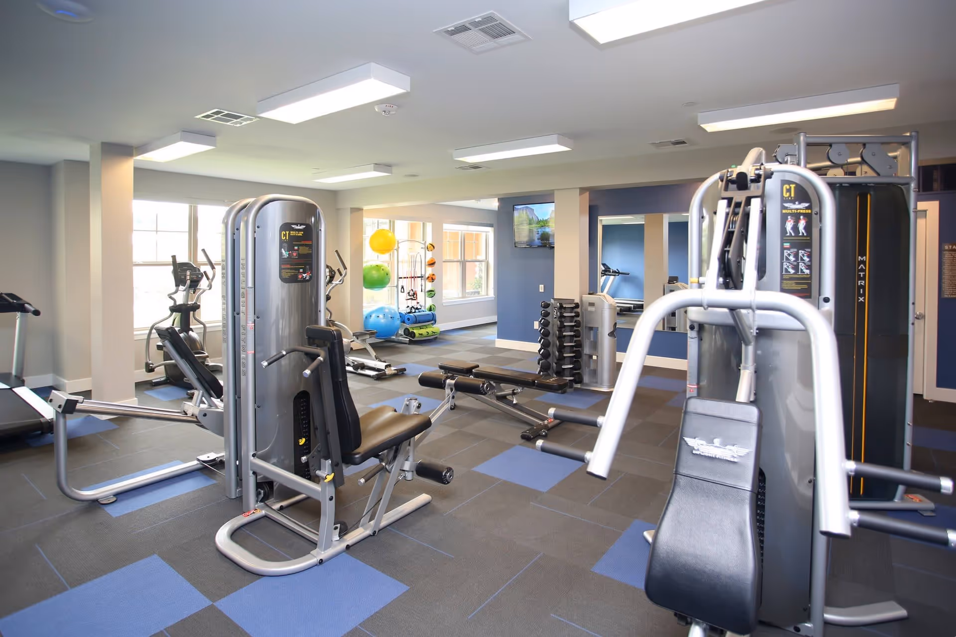 A well-lit fitness room with various exercise equipment including weight machines, dumbbells, treadmills, and colorful exercise balls near large windows.