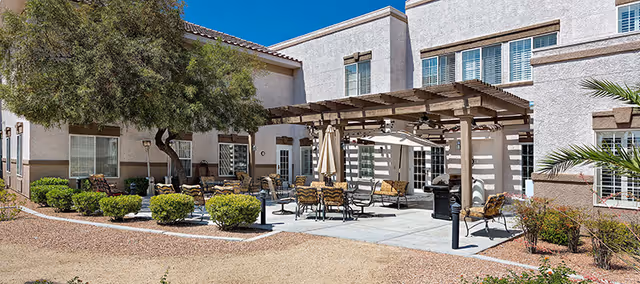 Sunlit outdoor courtyard with a wooden pergola, patio tables and chairs, umbrellas, and the facility's two-story building.