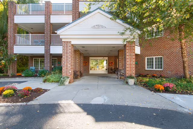 Entrance of a brick building with a covered driveway supported by brick columns. There are flower beds with colorful flowers on either side of the driveway and trees providing shade. The building has balconies with white railings and windows visible on the right side.
