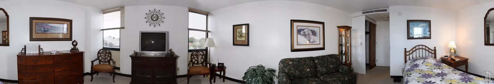 Panoramic view of a senior living studio with a bed, sofa, TV on a cabinet, dresser, chairs, and framed artwork.