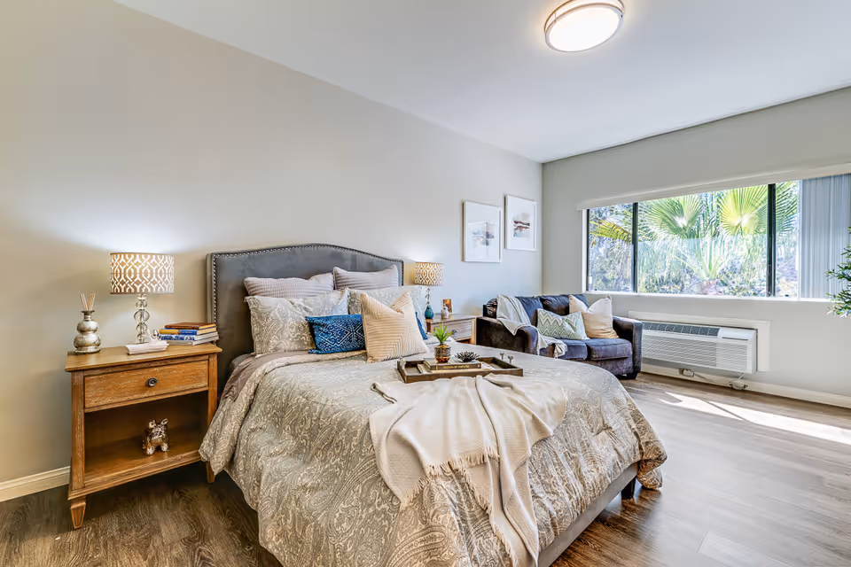 A bright and cozy bedroom in a senior living facility featuring a neatly made bed with multiple pillows and a throw blanket. There are two wooden nightstands with lamps and decorative items on either side of the bed. A comfortable armchair with cushions is placed near a large window that lets in natural light and offers a view of green palm trees outside. The room has light-colored walls and wood flooring.