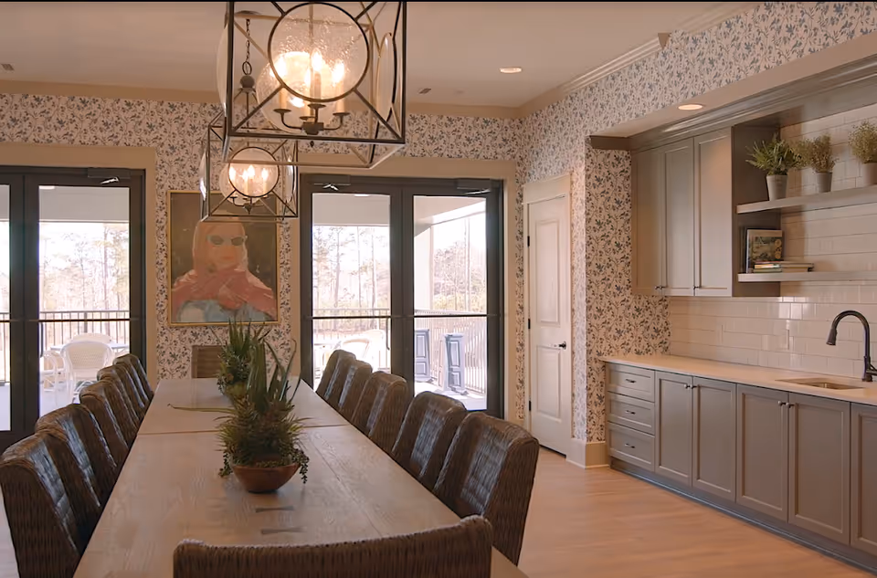 Long dining table with upholstered chairs under pendant lights in a room with built-in cabinets and glass doors leading to an outdoor balcony.