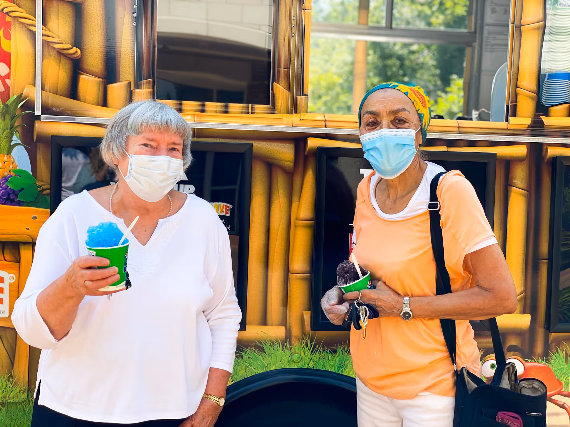 Two elderly women wearing face masks stand outdoors in front of a colorful food truck. One woman in a white top holds a blue shaved ice treat, while the other woman in an orange top and a colorful headscarf holds a purple shaved ice treat and carries a black bag.