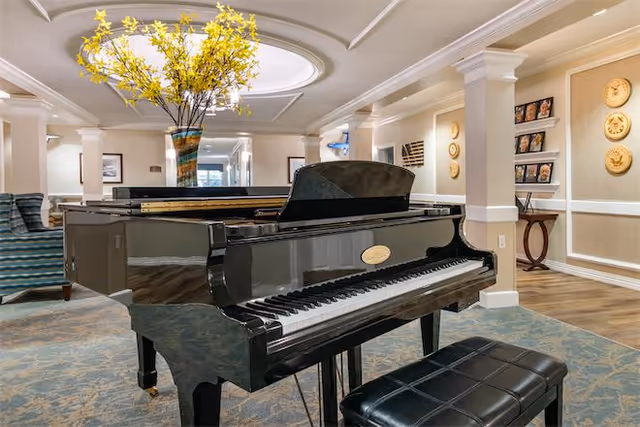 A polished black grand piano with a matching black cushioned bench in a well-lit room. Behind the piano, there is a large vase with yellow flowers on a table. The room features beige walls with white trim, decorative wall art, and comfortable seating including a striped armchair. The ceiling has a circular recessed light fixture.