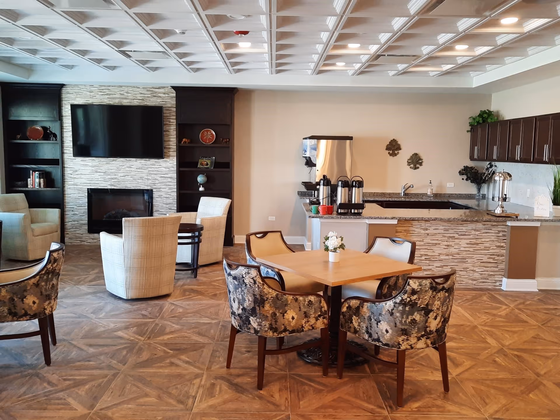 A cozy senior living common area featuring a seating arrangement with patterned and plain armchairs around a wooden table with a small flower vase. In the background, there is a modern fireplace with a mounted flat-screen TV above it, flanked by dark wooden shelves with decorative items. To the right, there is a kitchenette area with granite countertops, coffee dispensers, and cabinets, all under a coffered ceiling with recessed lighting.