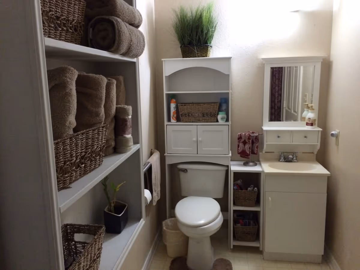 A small bathroom with a white toilet, a white cabinet above the toilet holding a basket and toiletries, a white sink with a mirror above it, and shelves on the left side stocked with rolled brown towels and baskets. There is a small plant on the top of the cabinet and another plant on the shelf. The bathroom has beige walls and a light-colored floor.