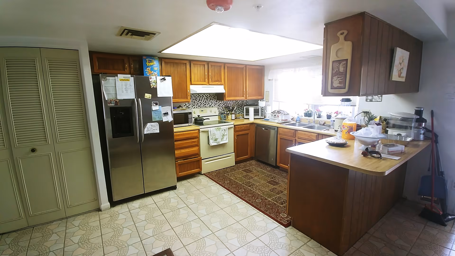 A kitchen with wooden cabinets, a stainless steel refrigerator covered with notes and magnets, a white stove with a towel hanging on the handle, a microwave, a dishwasher, and a countertop with various kitchen items. There is a window with blinds above the sink, a patterned rug on the tiled floor, and cleaning tools like a broom and dustpan in the corner.