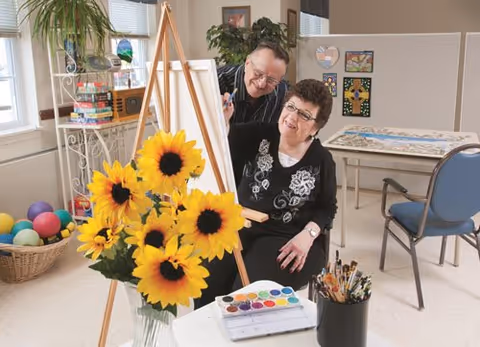 Two elderly individuals engaged in a painting activity in a bright room. One person is seated at an easel with a paint palette and brushes nearby, while the other person stands behind, smiling and interacting. A vase with large sunflowers is in the foreground, and the room contains colorful balls in a basket, artwork on the walls, and chairs.