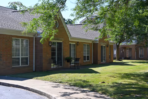 Exterior view of a single-story brick building with multiple windows and glass doors, surrounded by green grass and trees casting shadows on the lawn.