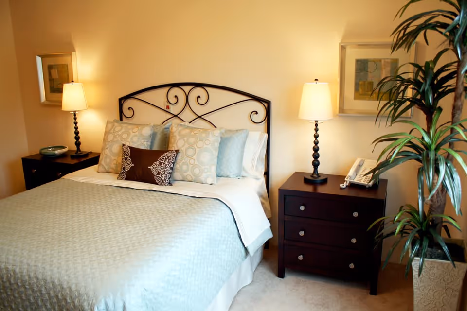 A neatly made bed with a decorative metal headboard, light blue quilt, and multiple pillows in a bedroom. On each side of the bed are dark wooden nightstands with lamps, a telephone on the right nightstand, framed artwork on the walls, and a tall potted plant on the right side.