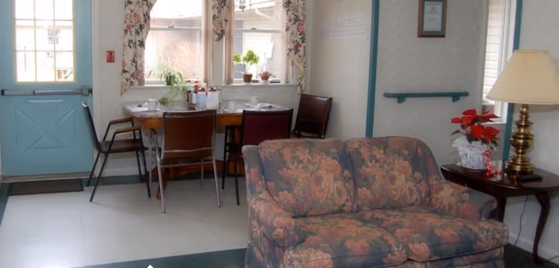 Small communal living area with a floral couch, side table and lamp, and a dining table with chairs by windows.