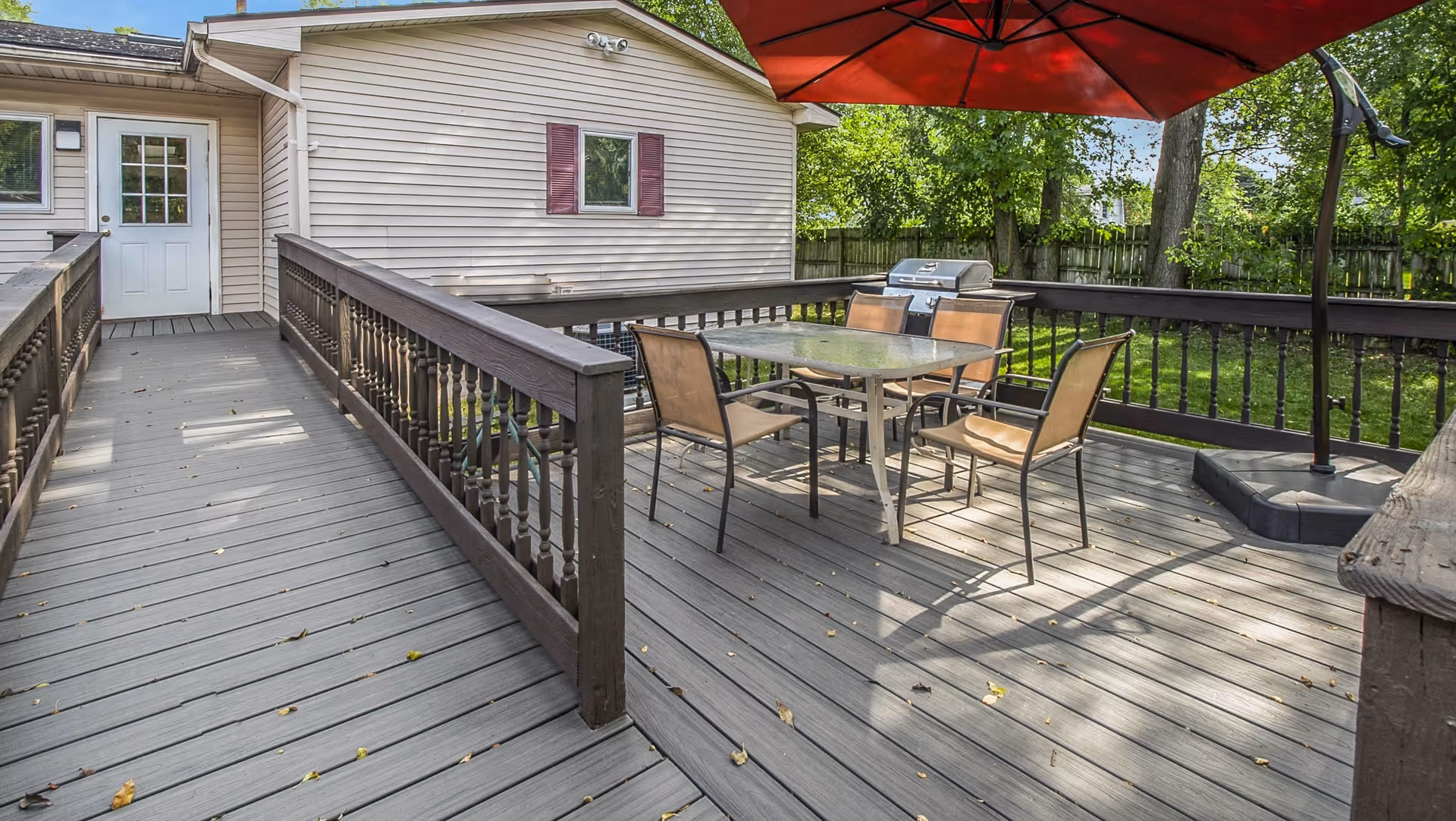 Outdoor wooden deck area with a glass-top table and four chairs under a large red umbrella. There is a barbecue grill in the corner and a white door leading into a building with beige siding and a small window with red shutters. The deck has wooden railings and is surrounded by green trees and a wooden fence.