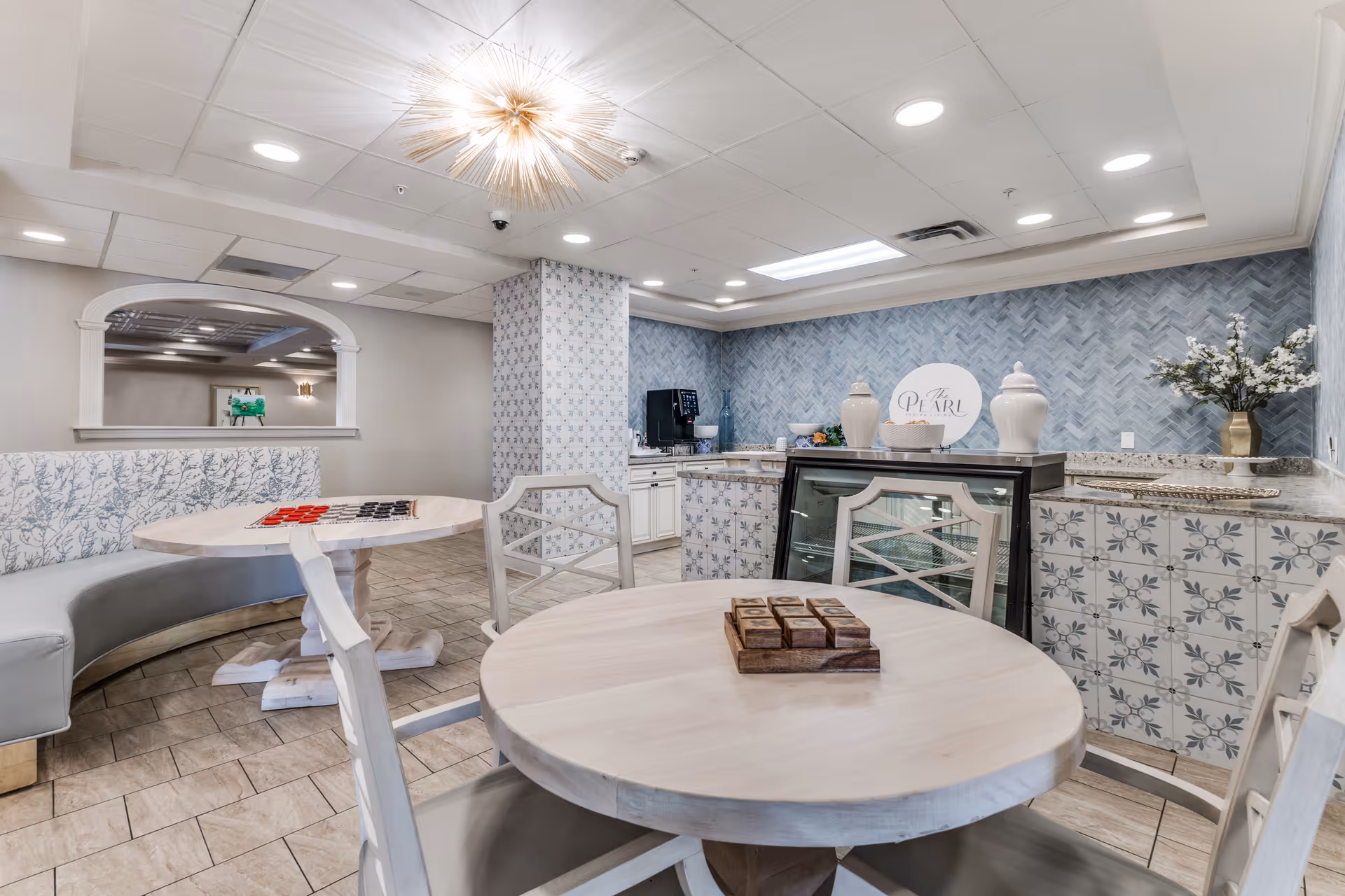 A bright and airy common area in a senior living facility featuring round wooden tables with chairs, a curved bench with patterned upholstery, a tiled counter with decorative vases, a coffee machine, and a sign that reads 'The Pearl'. The walls have a blue patterned design and the ceiling has recessed lighting and a modern chandelier.