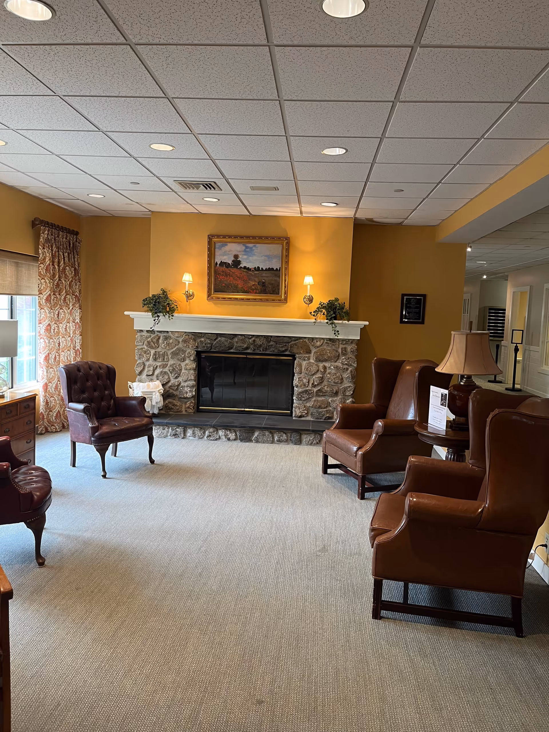 A cozy living room area with a stone fireplace centered on a mustard yellow wall. Above the fireplace is a framed painting flanked by two wall-mounted lamps. There are four leather armchairs arranged around the fireplace, two on the left and two on the right. A window with patterned curtains is on the left side, and a table with a lamp and informational papers is on the right. The ceiling has recessed lighting and a white drop ceiling.