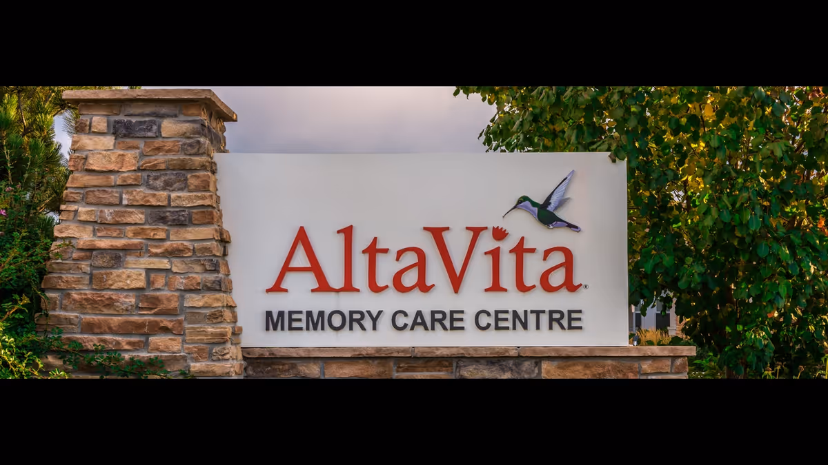 Stone and brick sign for AltaVita Memory Care Centre with a hummingbird graphic on the right side, surrounded by greenery and trees.