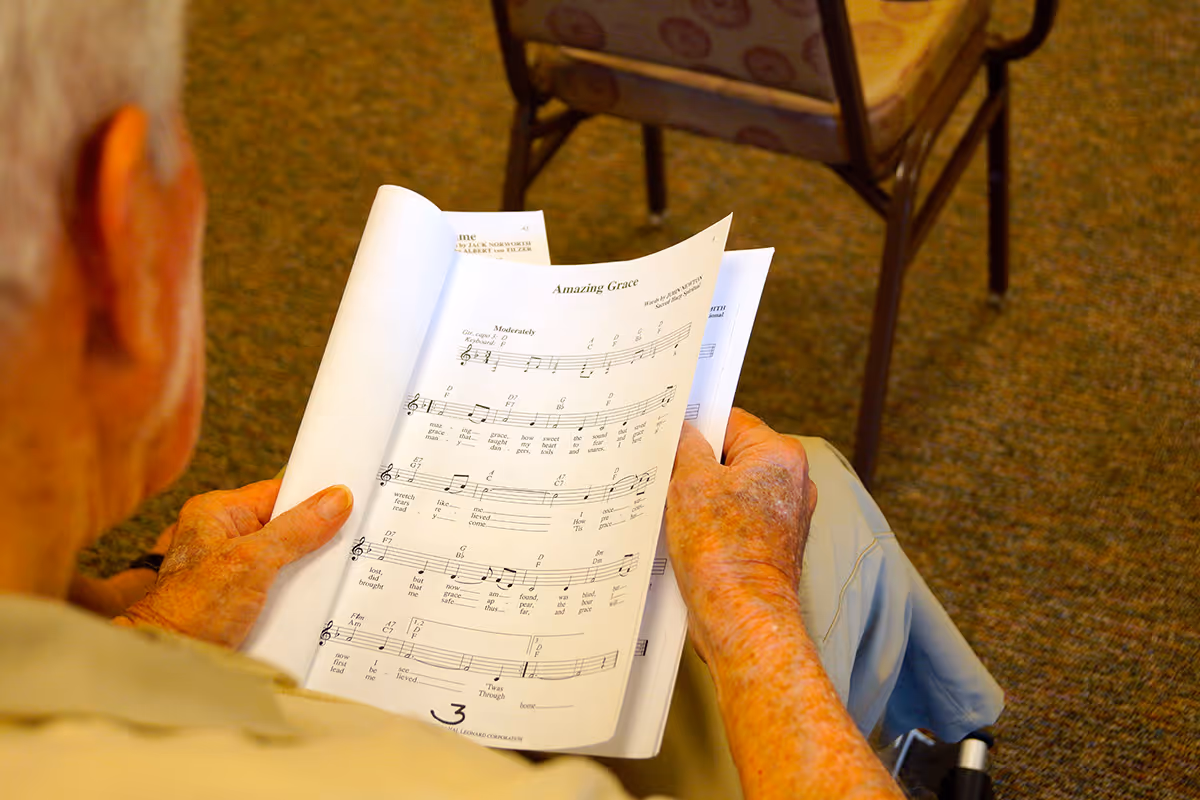 An elderly person holding and looking at sheet music for the song 'Amazing Grace' while seated on a chair in a room with carpeted floor and another chair visible in the background.