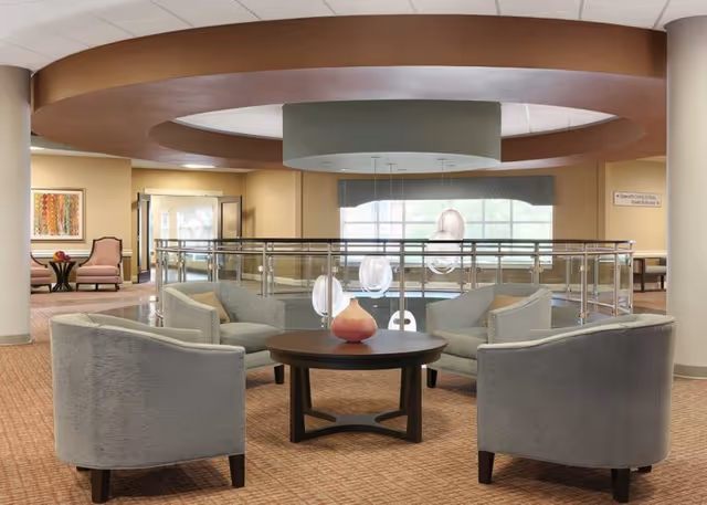 A spacious and well-lit common area with four gray upholstered armchairs arranged around a round wooden coffee table with a decorative vase. The area features a circular ceiling design with hanging pendant lights and large windows in the background. The space has beige walls and carpeted flooring, with additional seating and artwork visible in the background.