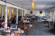 A spacious dining room with multiple tables and chairs arranged neatly. The tables are set with condiments and napkins. The room has carpeted flooring, large windows allowing natural light, and decorative hanging ornaments from the ceiling.