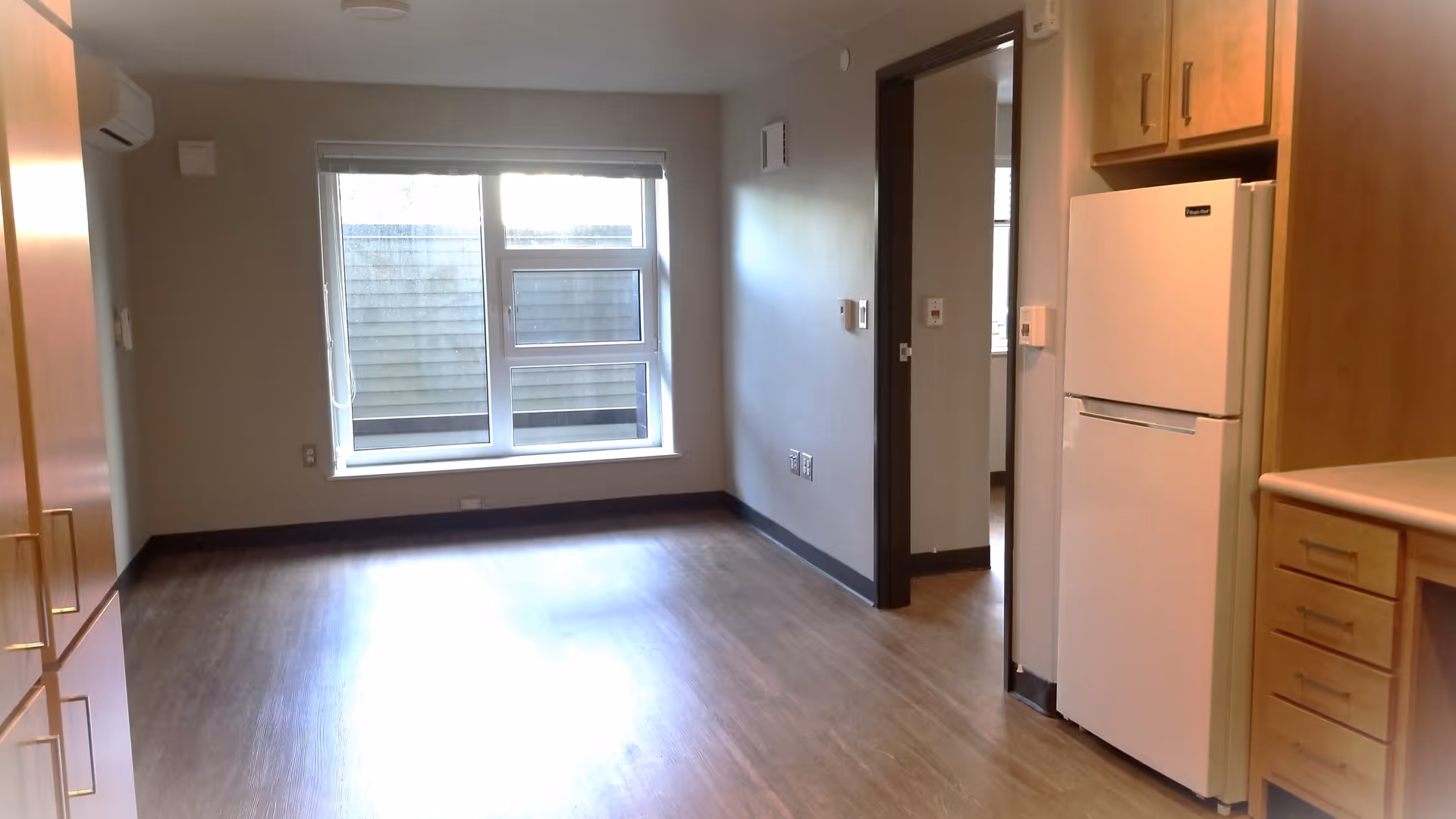 Empty studio-style living area with wood floors, a window at the far wall, and a kitchenette with a refrigerator and cabinets on the right.