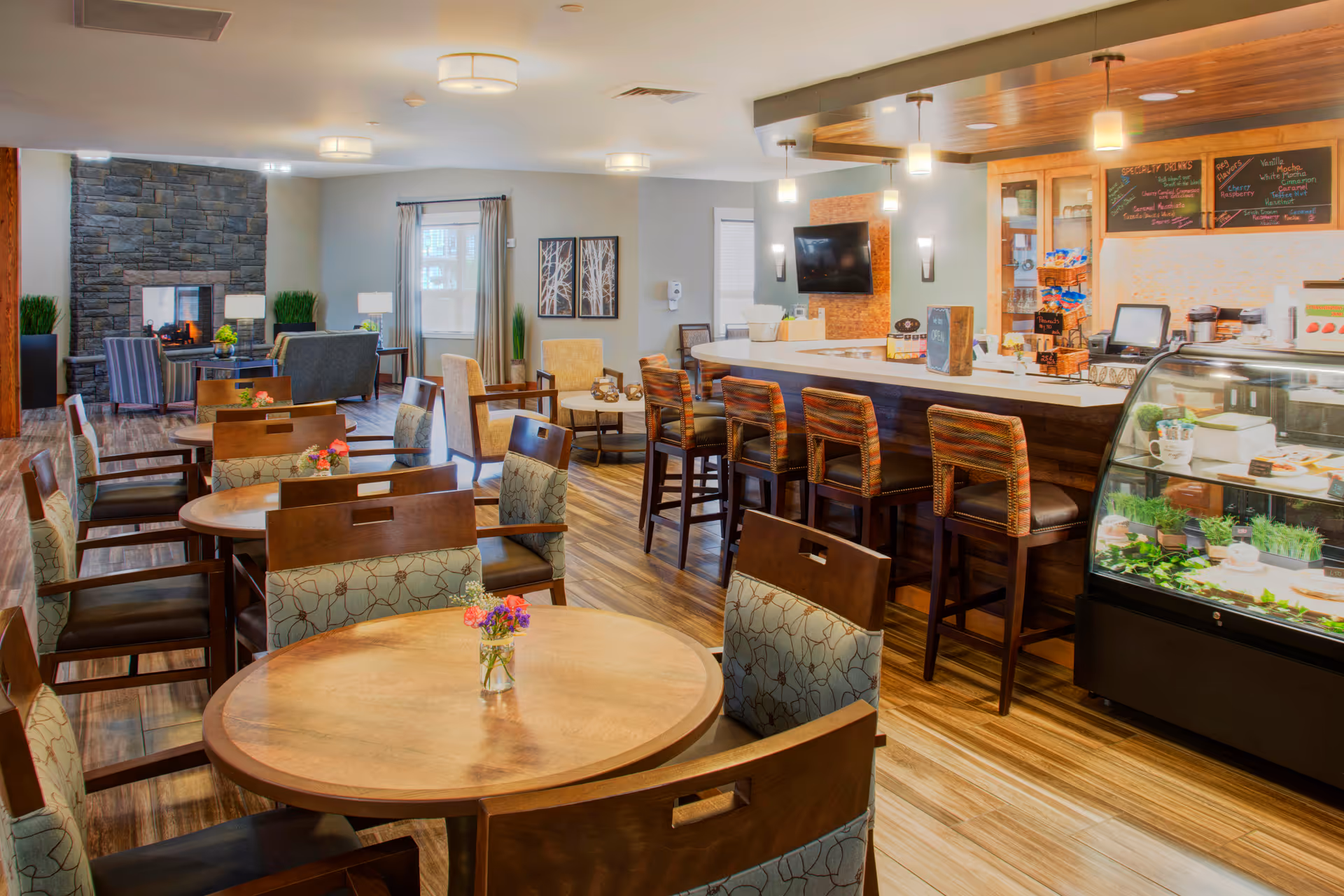 A cozy dining and lounge area in a senior living facility featuring round wooden tables with chairs, a stone fireplace in the background, a bar counter with high stools, and a display case with food items. The room has wooden flooring, soft lighting, and decorative plants.