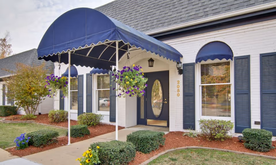 Exterior view of a white brick building with dark blue shutters and a matching blue awning over the entrance. The entrance has a glass door with an oval window, and hanging flower baskets with purple and yellow flowers are suspended from the awning. The building number 2050 is visible next to the door. There are neatly trimmed bushes and a mulched garden bed along the sidewalk leading to the entrance.